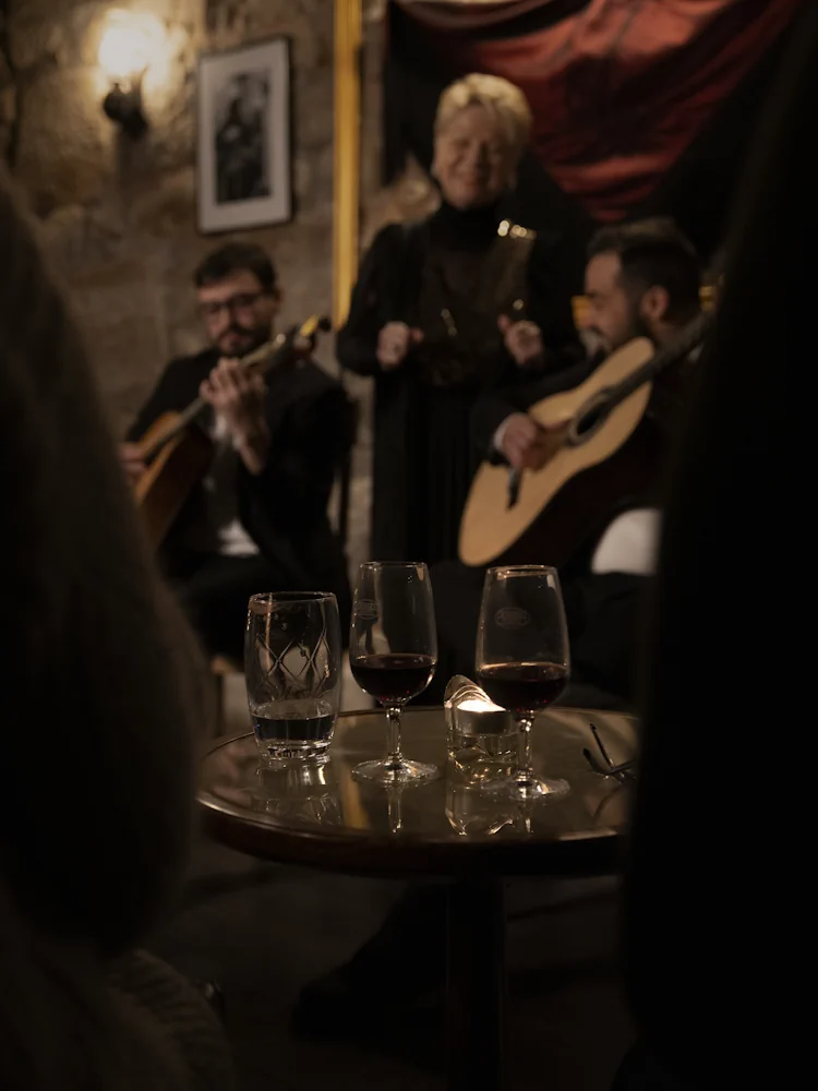 Cálices de vinho do Porto sobre mesa redonda, com dois guitarristas e uma fadista num concerto ao vivo no Porto, no Fado Maior do Porto