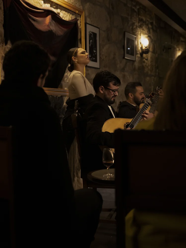 Fadista a cantar com dois guitarristas num concerto de Fado ao vivo no Porto, junto a paredes de granito e quadros antigos no Fado Maior do Porto